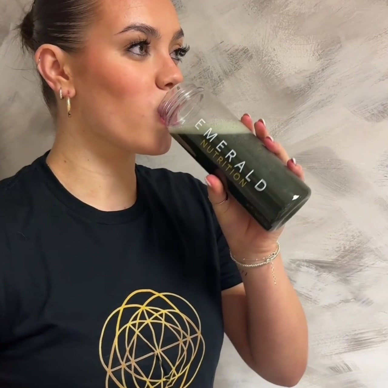 Woman drinking from a Emerald Nutrition bottle against a neutral background