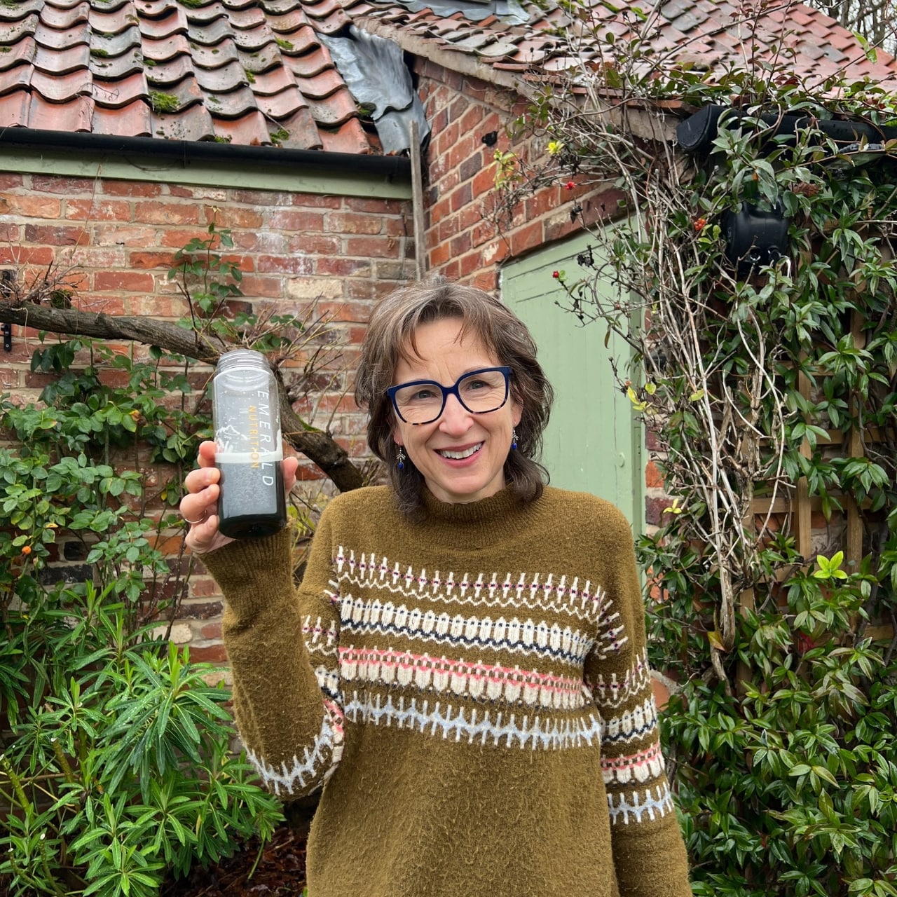 Woman holding a blender outdoors with a brick building and greenery in the background