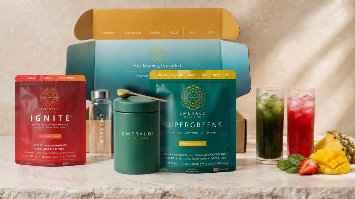 Emerald Wellness tea gift set featuring a teal and yellow box, a red Ignite pouch, a green container, a silver bottle, and two glasses of iced tea with fruit slices on a marble surface.