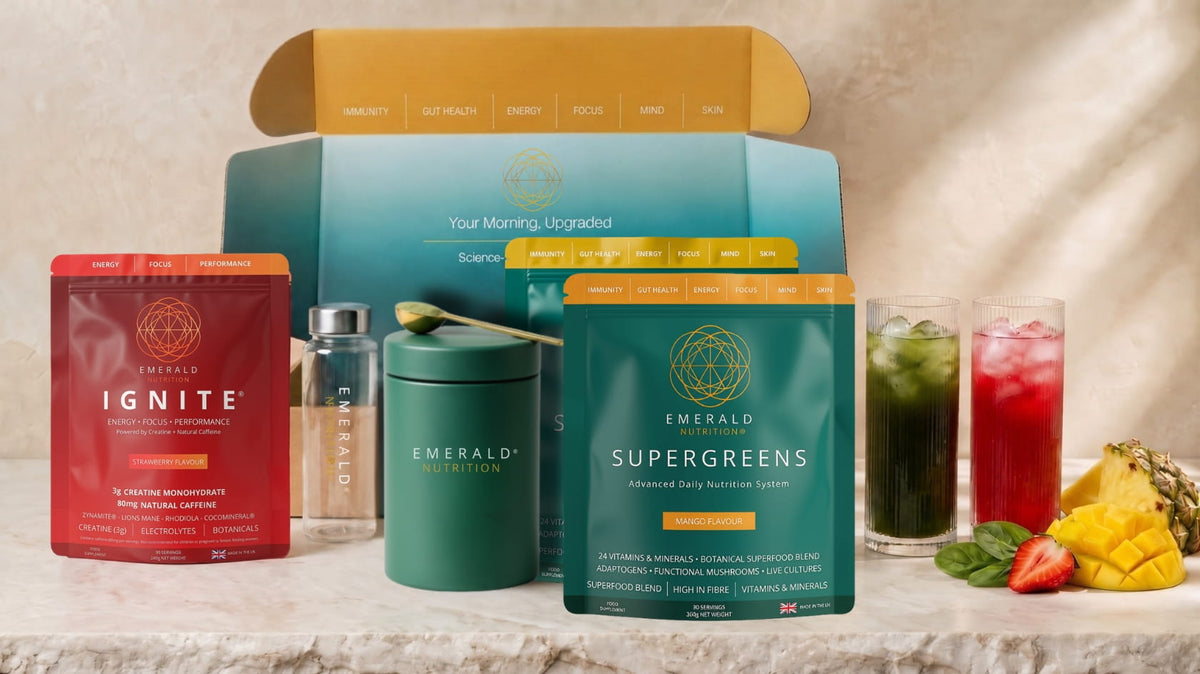 Emerald Organics wellness gift set featuring a red Ignite pouch, a green Emerald Supergreens pouch, a green canister, a small glass bottle, and an open teal and gold box, styled with two glasses of iced drink and fruit on a marble surface.